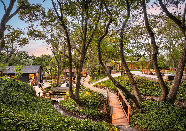 Image of a green landscape with cabins and a bridge over a small stream